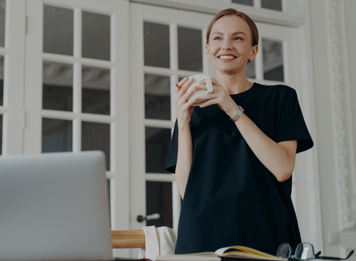 happy woman stands up holding a coffee mug at her desk, demonstrates what standing throughout the day does to your body