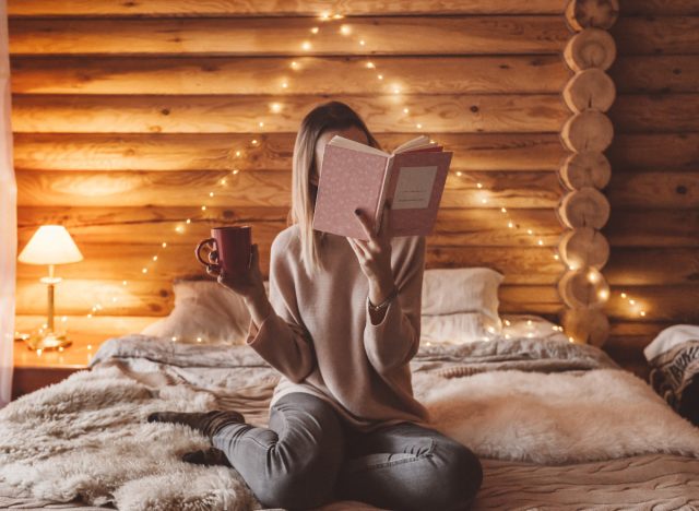 cozy winter cabin woman reading book and holding mug of coffee, concept of winter self-care hacks
