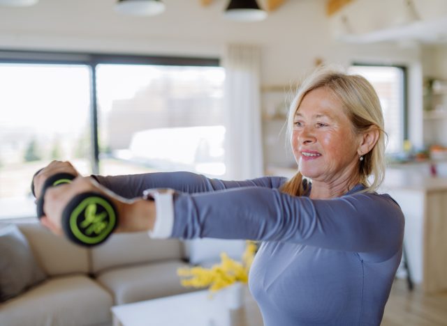 Fit senior woman at home showing body parts to exercise with dumbbells after age 60