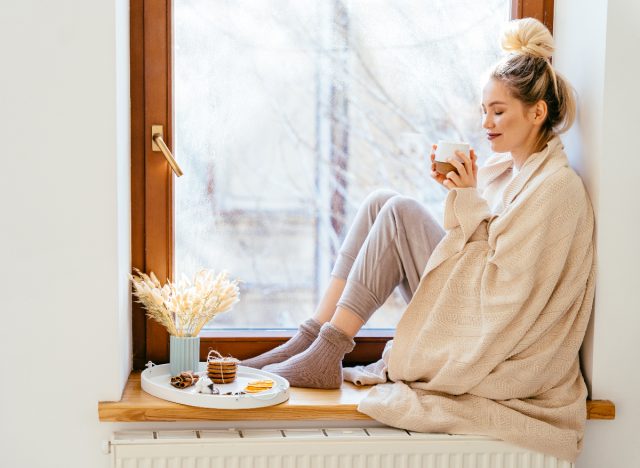 happy woman cozy sipping tea on windowsill in sunlight, demonstrating how to boost your mood in the winter