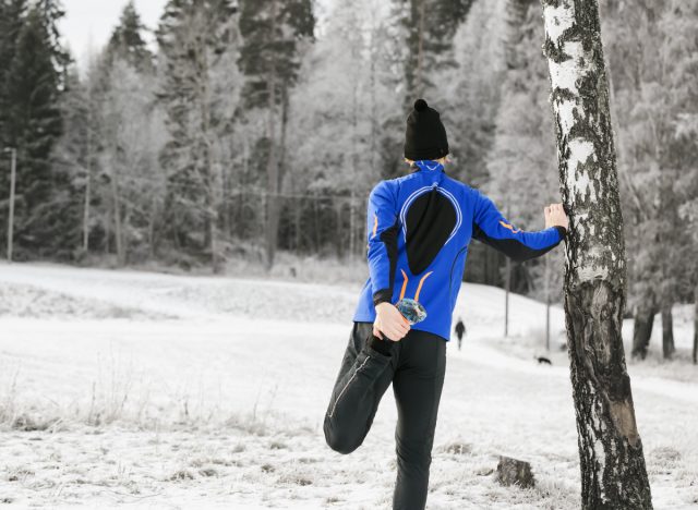 man in snow doing standing quad stretch against tree