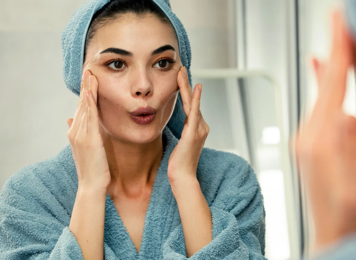 woman in bathrobe looking in mirror demonstrating face yoga exercises to lose face fat