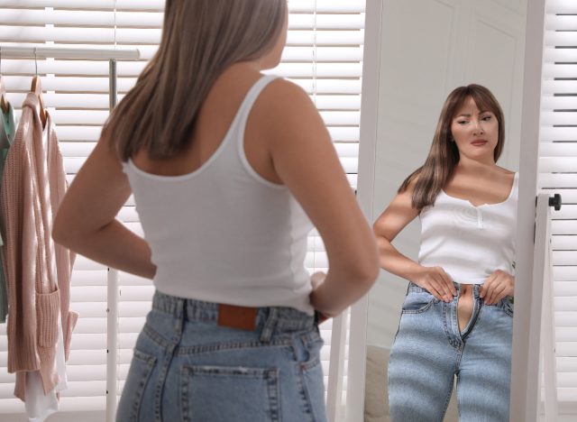 woman trying to button pants in mirror, concept of how to drop a size after weight gain