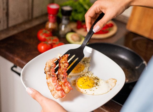 close-up woman's hands adding bacon to plate of eggs next to stove, concept of empty calorie foods that make you gain weight