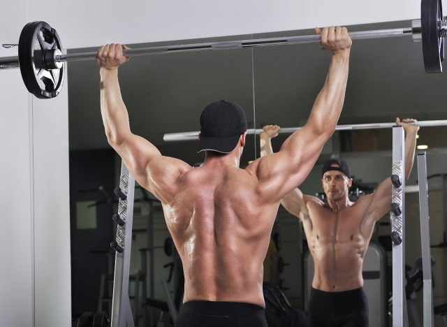 muscular man doing barbell press exercises in front of gym mirror, demonstrating how to build your 3D shoulders