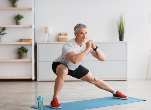 mature man at home doing couch potato workout side lunges