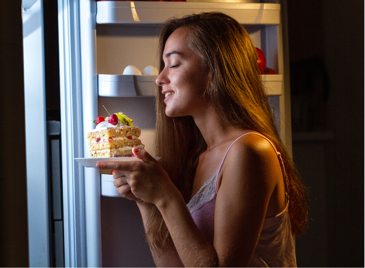 woman in PJs holding plate of cake by the fridge, concept of the worst late-night foods for weight loss