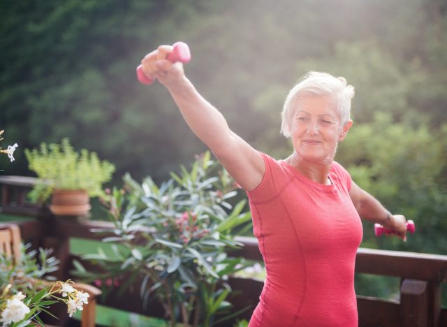 older woman holding dumbbells doing exercise outdoors on her deck