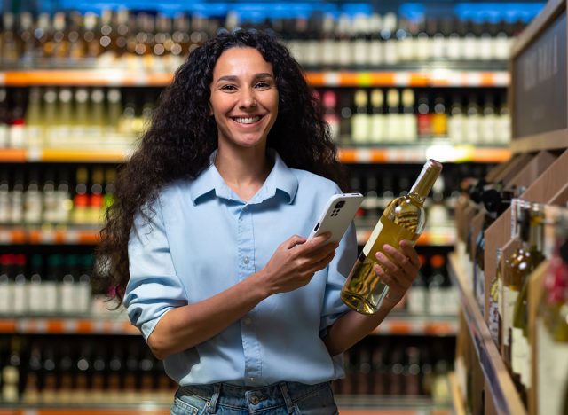 woman shopper in supermarket, using app on phone to scan labels and rate wine.