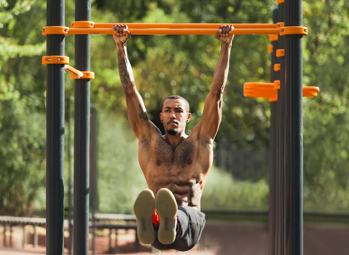 ripped, fit man doing leg raises on pull-up bar outdoors as part of upper-body workout for shredded torso