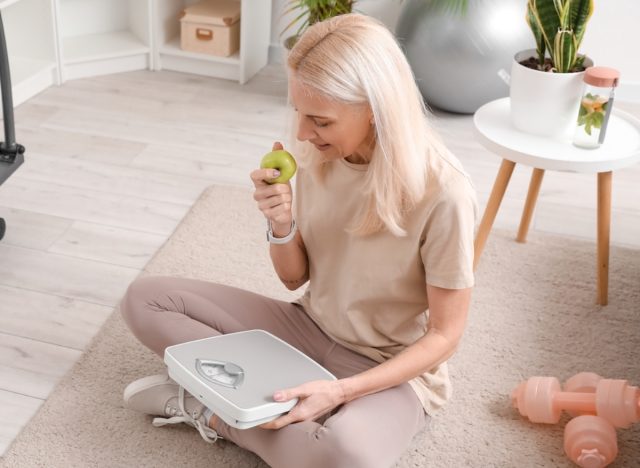 mature woman eating apple while holding scale, concept of eating habits for rapid weight loss after 50