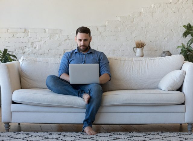 The 6 fitness habits that destroy your legs before you turn 40 = man sitting on couch typing on laptop