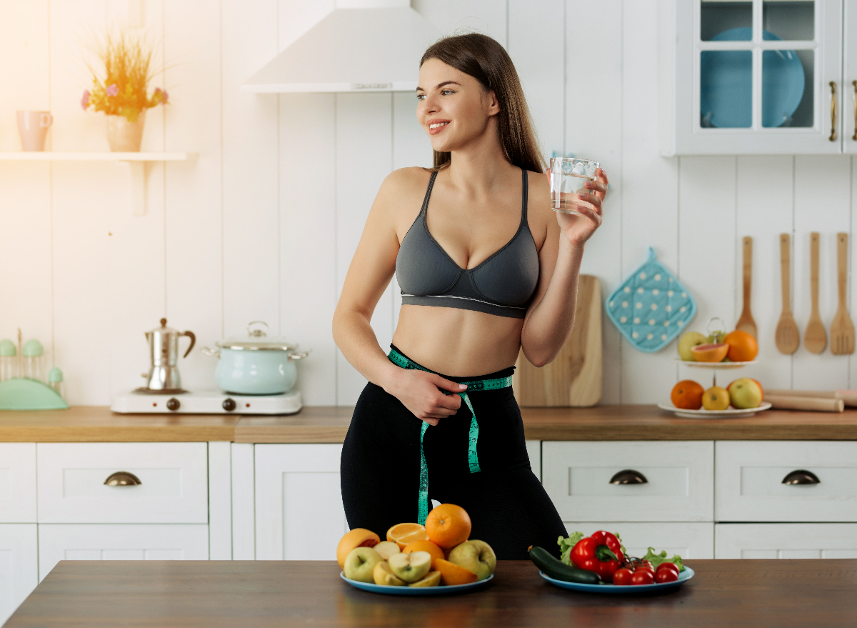 woman measuring waist, holding glass of water in kitchen, concept of ways to lose belly fat