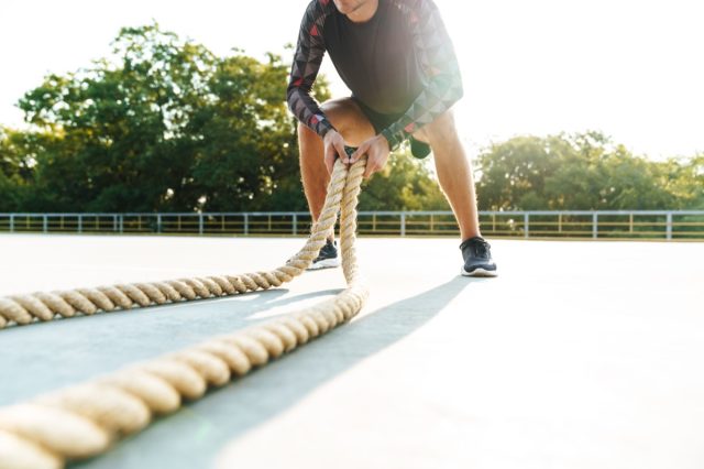 Cropped photo of athletic man in sportswear doing workout with battle ropes at playground outdoors