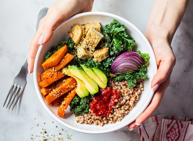 Woman hands eating vegan salad of baked vegetables, avocado, tofu and buckwheat buddha bowl, top view