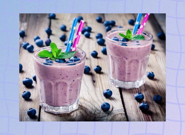 blueberry smoothies in two glasses with colorful straws surrounded by fresh blueberries on wood table