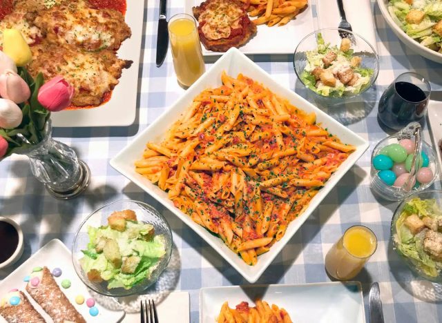 buca di beppo easter meal with pasta, salad, cannolis, chicken parmesan, mimosas, a glass of red wine, and bouquet of tulips on a light blue patterned tablecloth