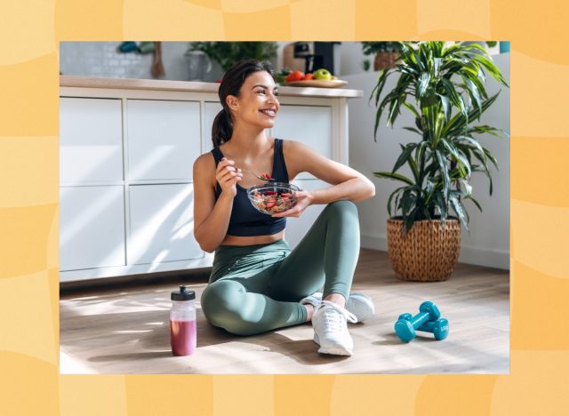 happy woman in green athletic set sitting on kitchen floor eating fiber cereal next to smoothie and dumbbells