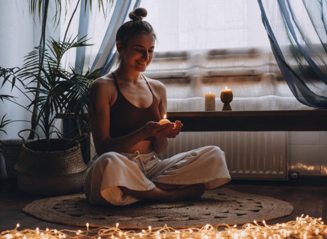 happy woman holding a candle and meditating in her living room, surrounded by string lights