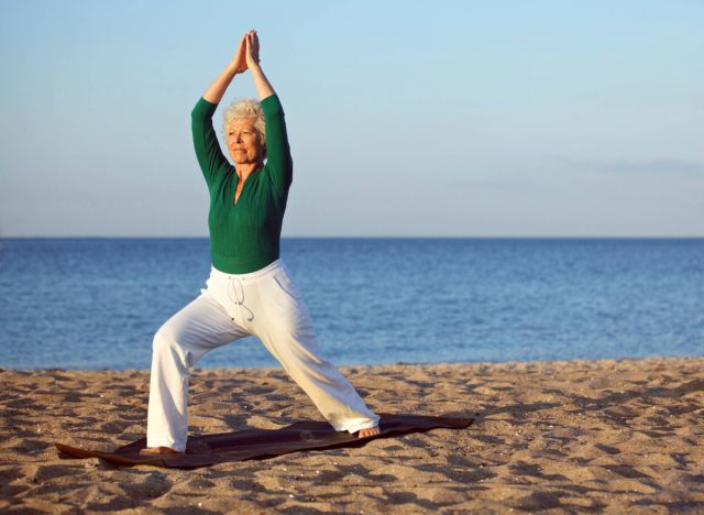 mature woman in green long-sleeve top and white pants doing yoga at golden hour on the beach