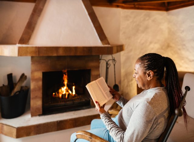 mature woman sitting in chair reading book by fireplace