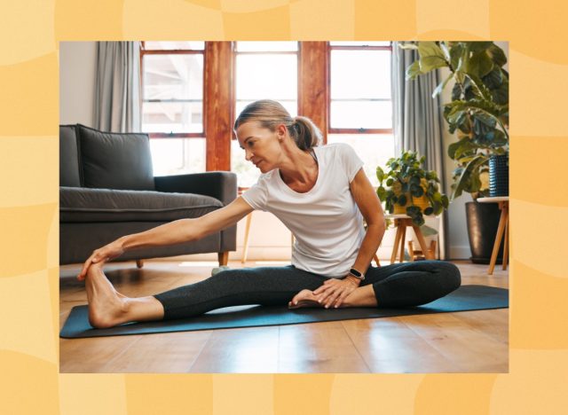 mature woman wearing white t-shirt and black leggings stretching her legs on yoga mat in bright living room in front of window
