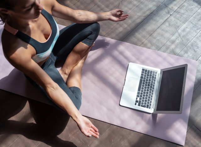 woman in yoga apparel doing meditation at home in front of laptop on mat