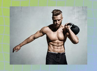 muscular man wearing gym shorts holding a kettlebell by his shoulder in front of gray backdrop