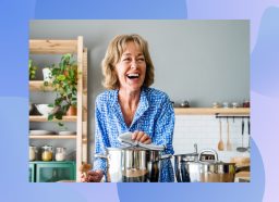 happy senior woman wearing blue button-down shirt laughing and cooking a healthy meal or soup in bright kitchen