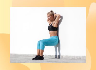 blonde woman wearing a black sports bra, bright blue leggings, and black sneakers doing wall squats outdoors against bright white wall