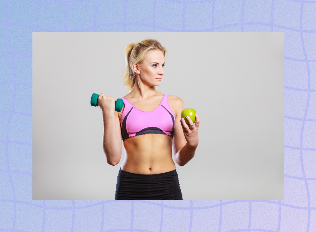 fit blonde woman in sports bra holding dumbbell and green apple in front of gray backdrop