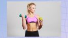 fit blonde woman in sports bra holding dumbbell and green apple in front of gray backdrop