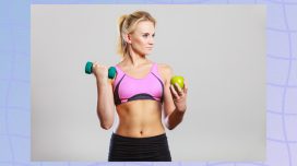 fit blonde woman in sports bra holding dumbbell and green apple in front of gray backdrop
