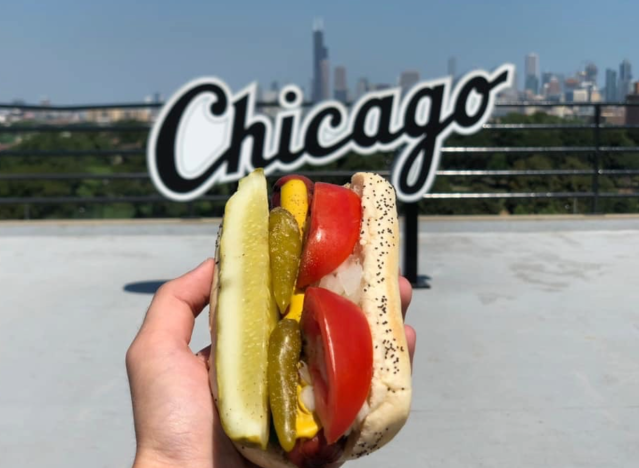 a hand holding a chicago style hot dog in front of chicago sign.