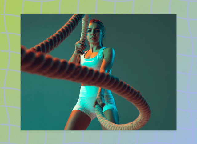 fit woman in white biker shorts and sports bra doing battle ropes exercise in front of green backdrop