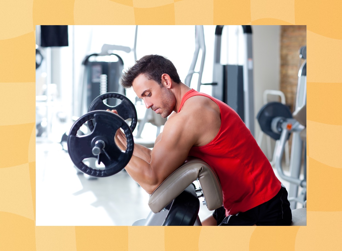 fit, focused man in red tank top and black gym shorts lifting barbell in bright gym