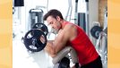 fit, focused man in red tank top and black gym shorts lifting barbell in bright gym