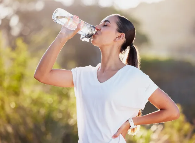 happy brunette woman in white t-shirt drinking water bottle outdoors on sunny day after workout