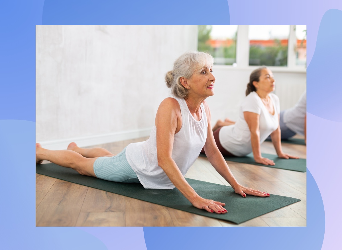 mature happy grey-haired woman doing cobra pose on dark green yoga mat in bright yoga classroom alongside other senior classmates