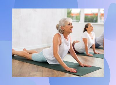 mature happy grey-haired woman doing cobra pose on dark green yoga mat in bright yoga classroom alongside other senior classmates