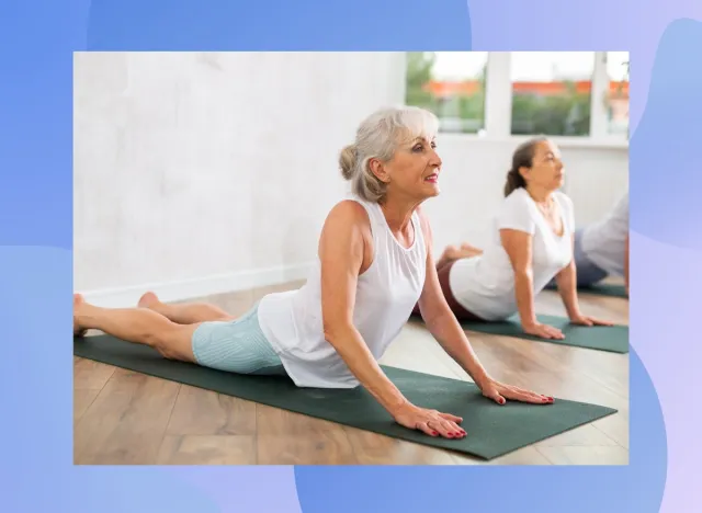 mature happy grey-haired woman doing cobra pose on dark green yoga mat in bright yoga classroom alongside other senior classmates