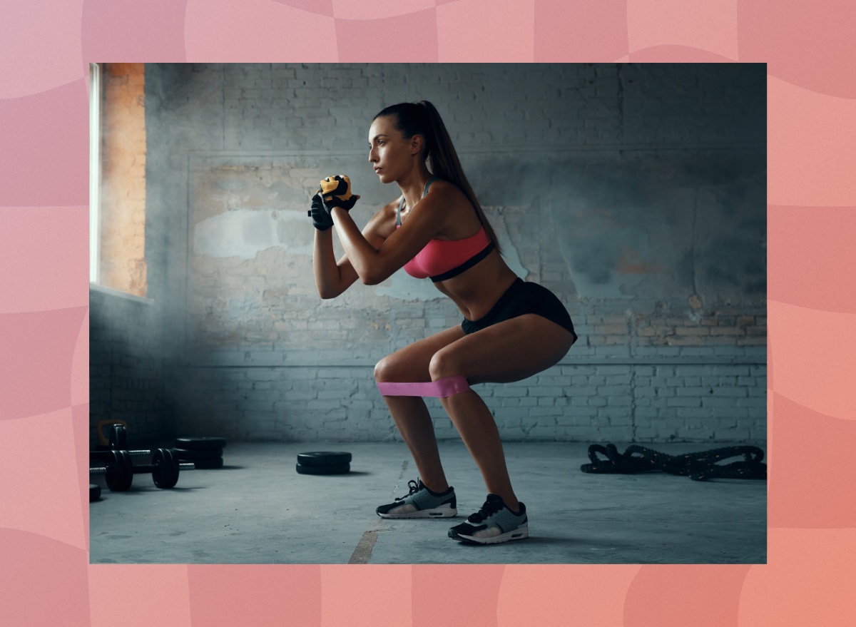 fit, focused brunette woman in gym shorts and pink sports bra doing resistance band squats at the gym