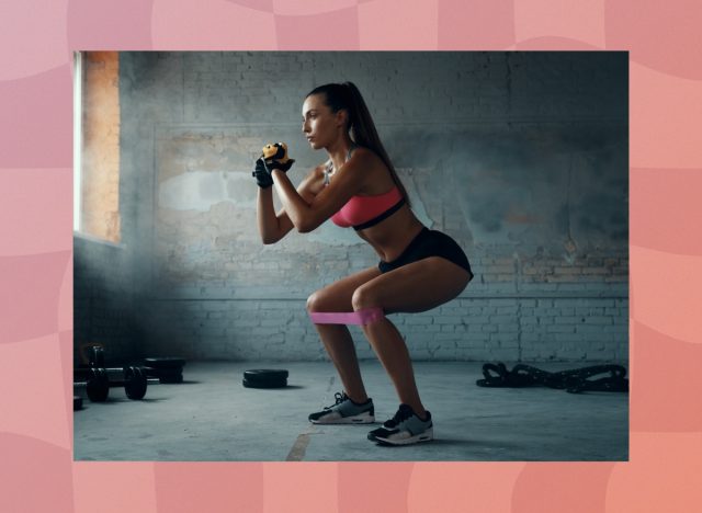 fit, focused brunette woman in gym shorts and pink sports bra doing resistance band squats at the gym