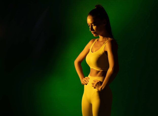 woman in yellow sports bra and leggings doing stomach vacuum exercise in front of green backdrop