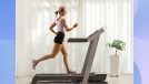 happy blonde woman in pink tank top and black biker shorts running on treadmill at home in bright living space in front of curtain window