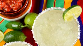 Margaritas and salsa on a colorful table cloth, with limes, and peppers.