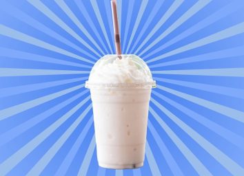 A classic vanilla milkshake in a see-through plastic cup with a straw set against a vibrant blue background