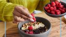 woman putting raspberries on top of a bowl of yogurt with chocolate chips and bananas