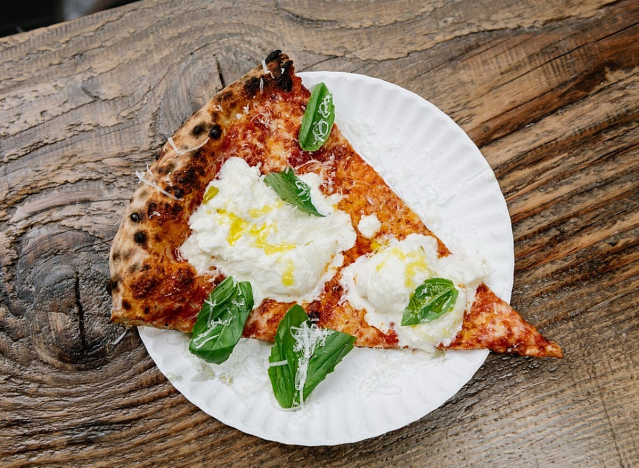 a slice of pizza with burratta and fresh basil on a wood table.