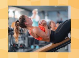 focuses brunette woman in orange sports bra and black workout leggings doing dumbbell sit-ups